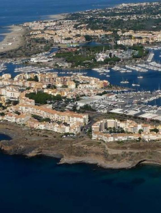 Vue aérienne du Cap d’Agde montrant le port de plaisance, les immeubles en bord de mer, la plage et le littoral méditerranéen.